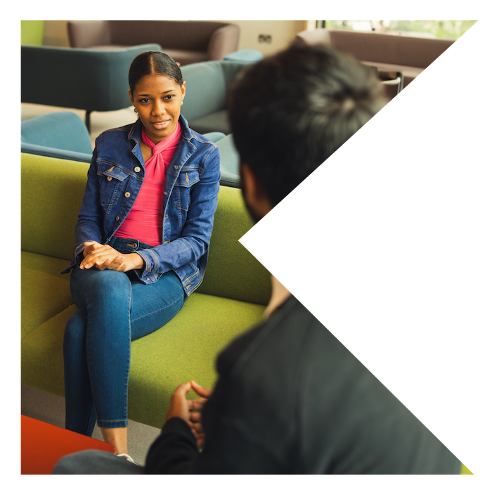 Black person wearing a pink top and denim jacket sitting on a green sofa speaking to someone