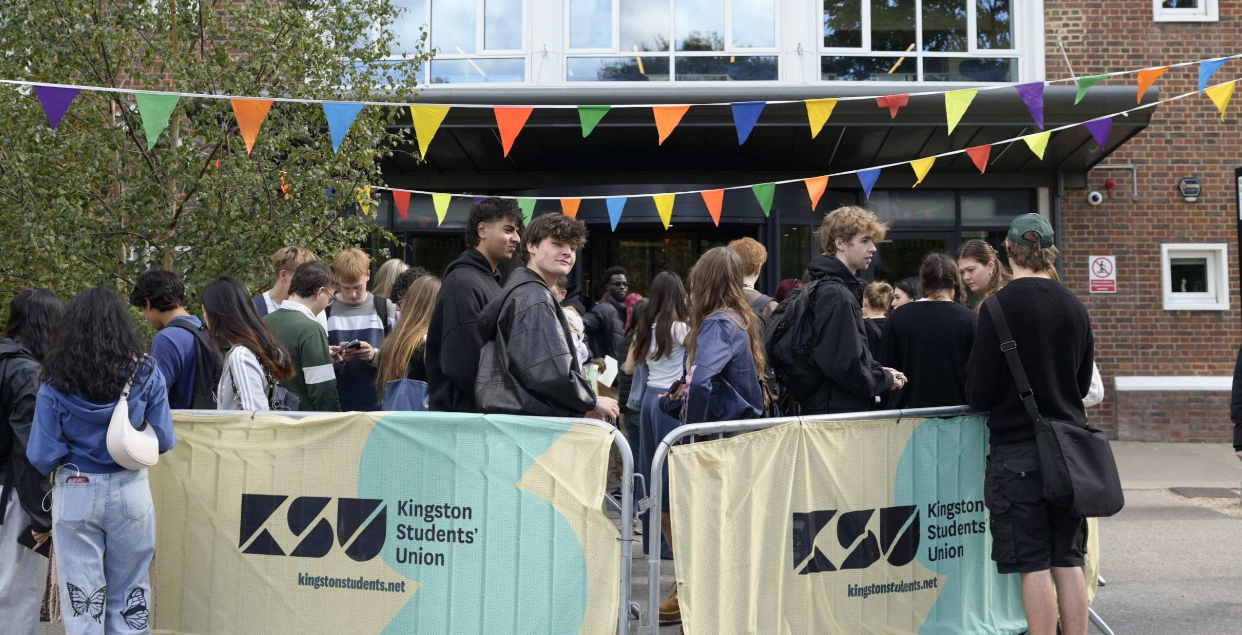 A group of students in a queue. The images have two barriers with mesh covers promoting Kingston Students' Union and bunting.